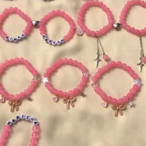Pink translucent bead bracelets with white letter beads spelling names, heart halves, bow, star and butterfly charms on cream fabric