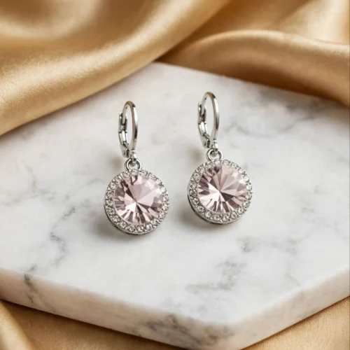 Round pale pink crystal drop earrings with clear rhinestone halo on silver-tone huggie hoops on a marble slab