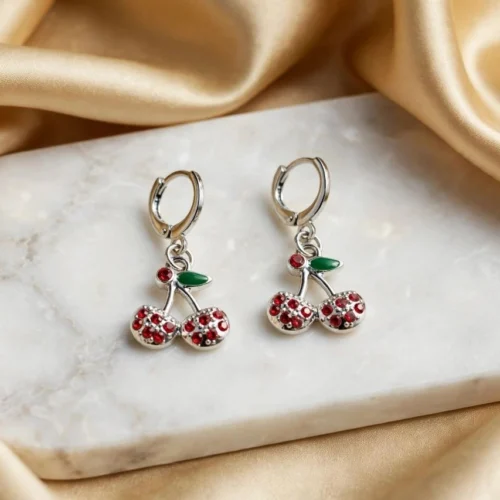 Silver-tone cherry earrings with red rhinestones and green enamel leaf on huggie hoops on a white marble board
