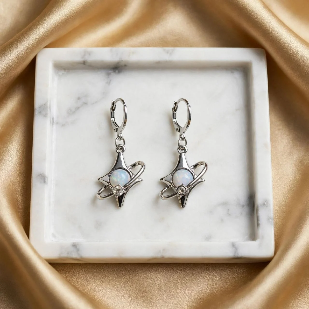 Silver-tone four-point star earrings with orbit ring, white opal-look stone and rhinestone on huggie hoops in a square marble tray