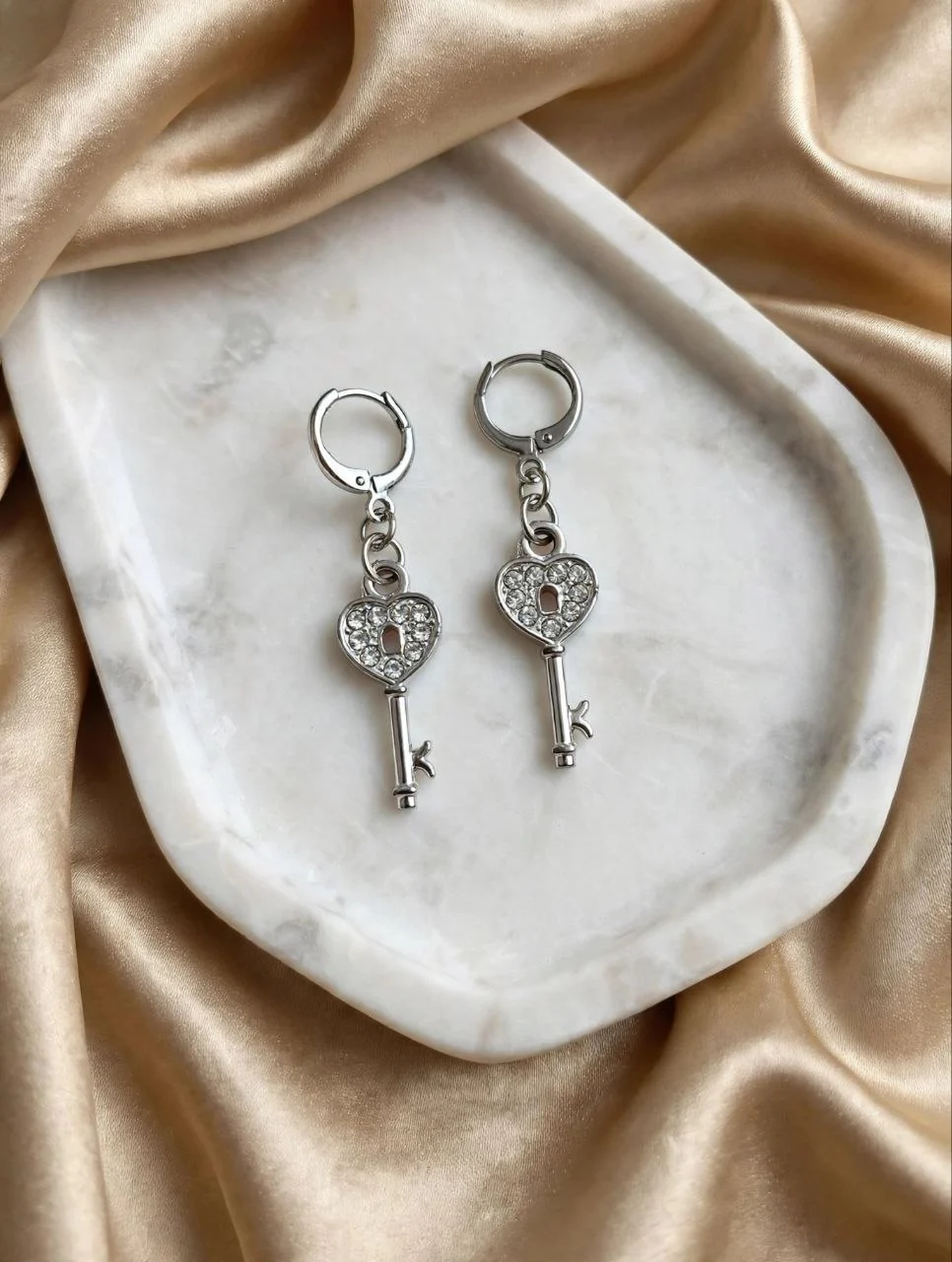 Silver-tone heart key earrings with clear rhinestones and keyhole on huggie hoops on a white marble tray