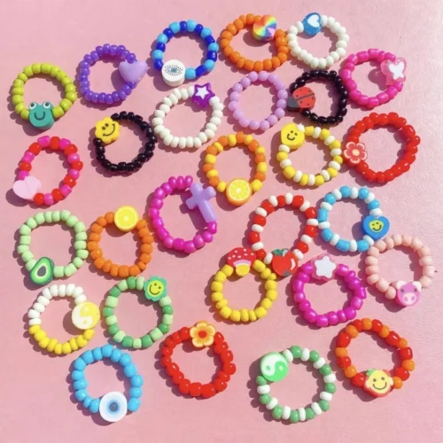 Pile of colourful seed bead stretch rings with smiley, fruit, flower and evil eye charm beads on a pink background
